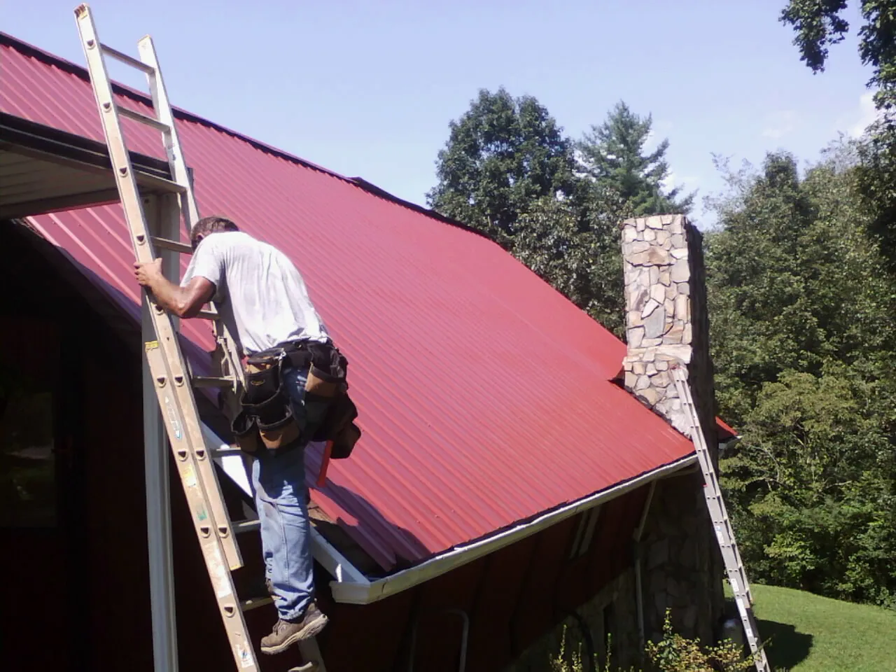 Roofer on a ladder beside a red metal roof and stone chimney — Soffit & Fascia Repair by Roofing Brothers in Arion